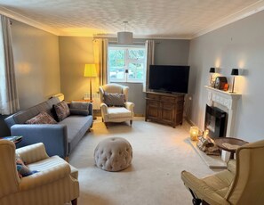 A living room with a sofa, armchairs, a round ottoman, a television, a fireplace, and a window with curtains. - No 1 The Martins (Reydon)