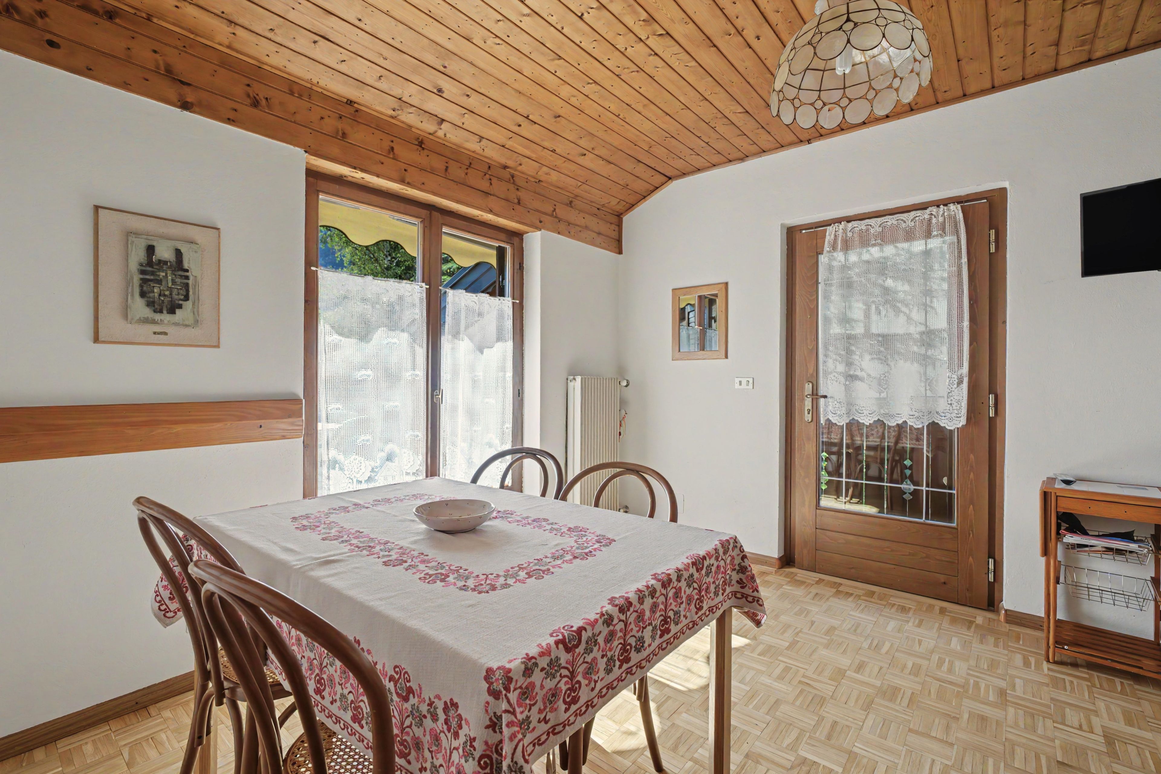 A dining area with a table set for two, wooden chairs, a ceiling light fixture, and a door with lace curtains.