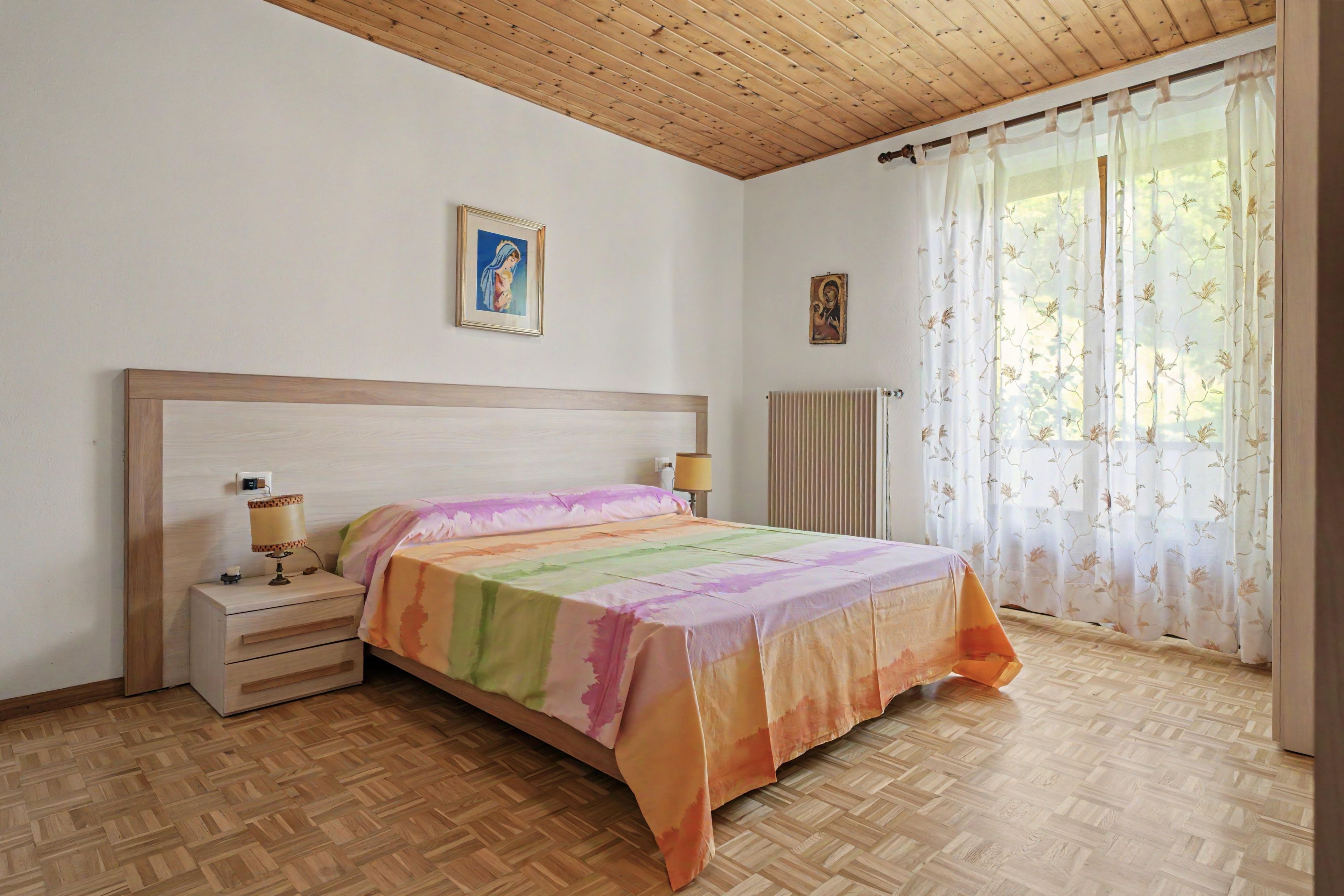 Ein Schlafzimmer mit Holzboden, einem Bett mit farbenfroher Bettdecke, zwei Nachttischen mit Lampen, einem Heizkörper und einem Fenster mit blumigen Vorhängen.