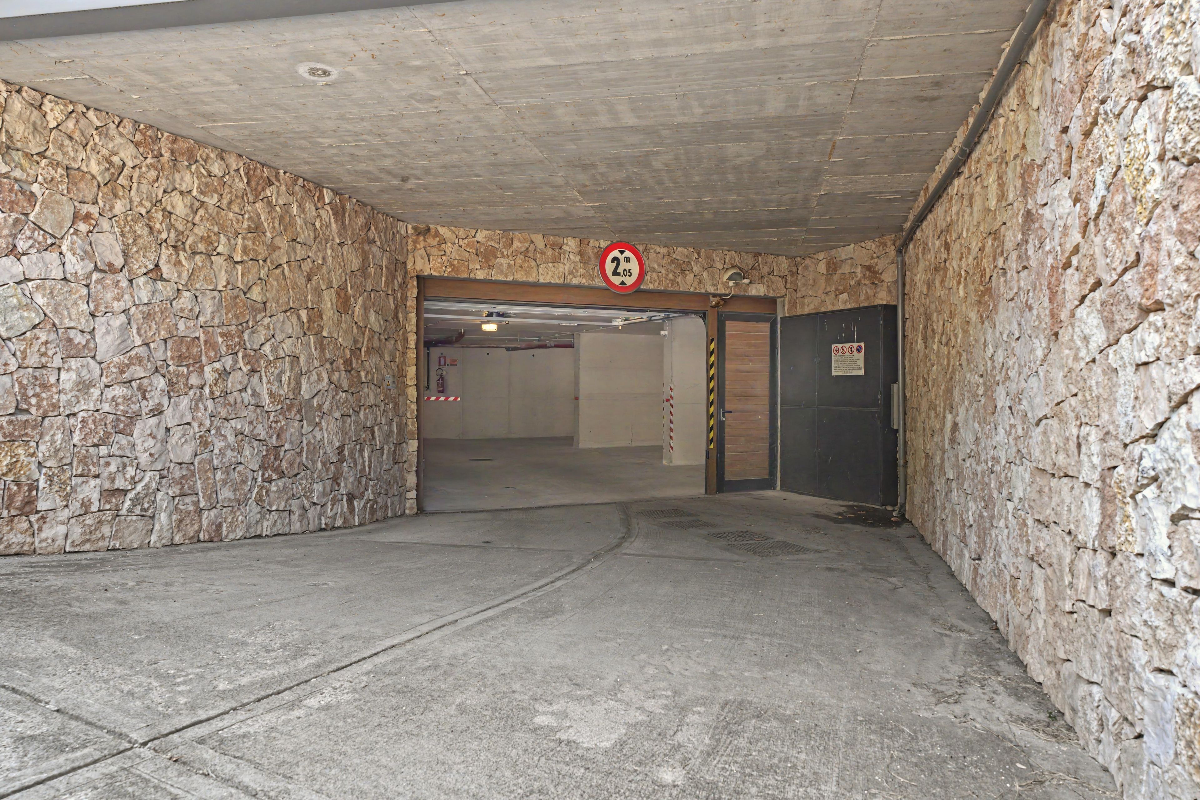 L’entrée d’un parking, avec des murs en pierre, un panneau indiquant la hauteur maximale autorisée et une porte de garage fermée.