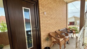 Ein Balkon mit einer dunkelholzigen Tür, Korbmöbeln, Topfpflanzen und einer Ziegelwand.
