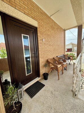 Ein Balkon mit einer dunkelholzigen Tür, Korbmöbeln, Topfpflanzen und einer Ziegelwand.