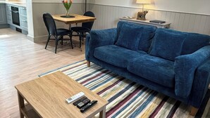 Ein Wohnzimmer mit blauem Sofa, einem Holztisch, einem Streifen Teppich, einem Esstisch mit Stühlen, einer Küchenzeile und Bildern an der Wand.