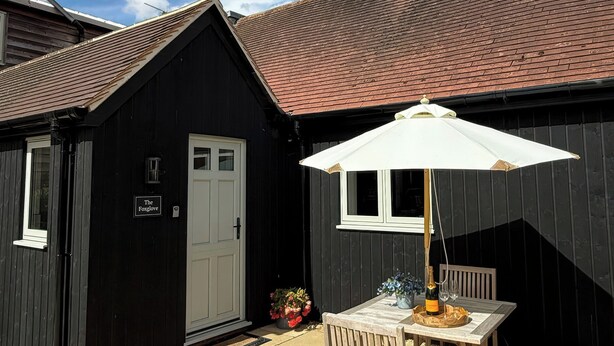 Ein schwarzes Holzgebäude mit einer weißen Tür und Fenstern, eine Terrasse mit Tisch und Stühlen sowie ein weißer Sonnenschirm.