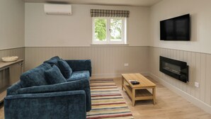 Una sala de estar con un sofá azul, una mesa de centro de madera, un televisor montado en la pared y una ventana con un adornos a cuadros.