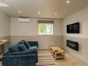 Ein Wohnzimmer mit einem blauen Sofa, einem Holztisch, einem an der Wand befestigten Fernseher und einem Fenster mit kariertem Vorhang.