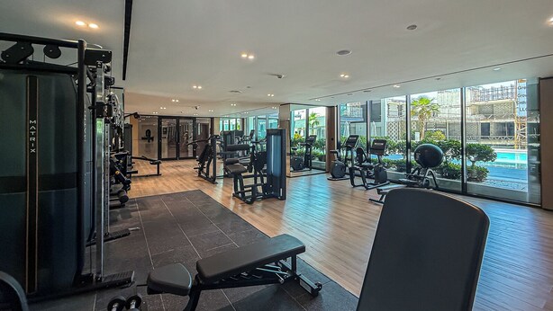 A gym with various exercise equipment, a bench, and a large window offering a view of the outside. - Elegant 3BR Stay in JVC near Circle Mall (دبي)