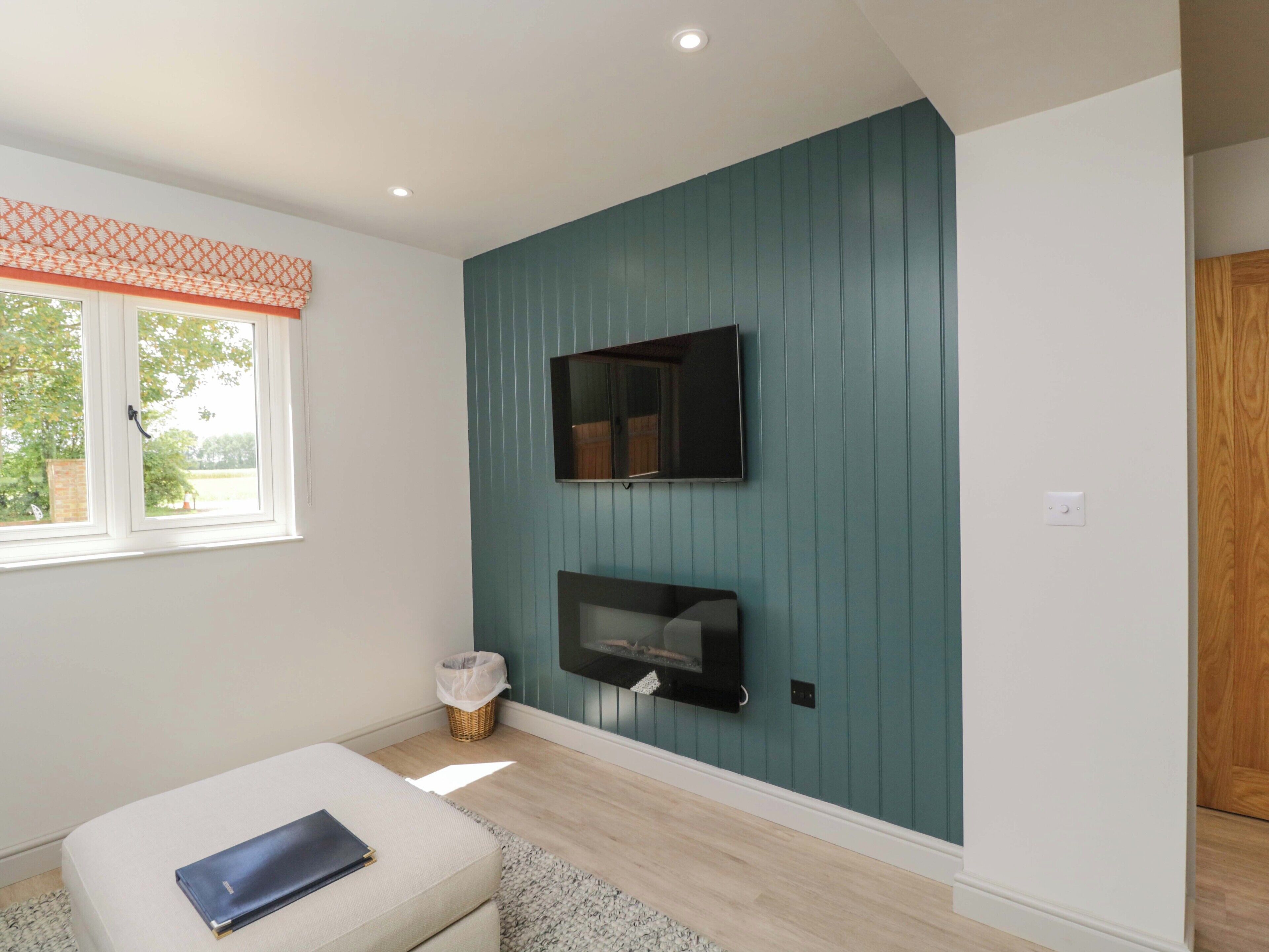A room with a television mounted on a teal wall, a window with a patterned valance, a white bench, and a wicker basket on the floor.