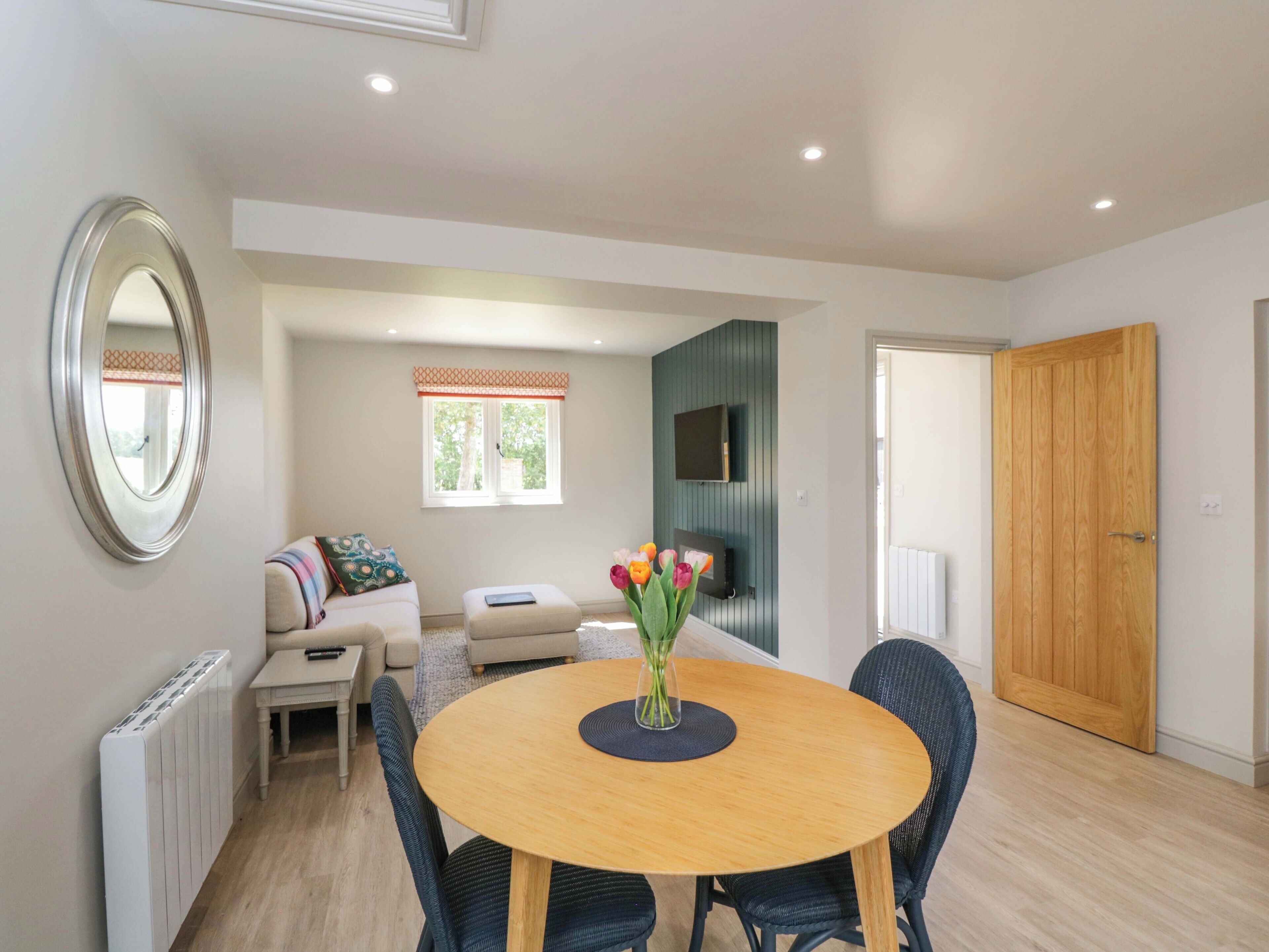 A modern living room with a round wooden dining table, black chairs, a sofa, a TV mounted on the wall, and a wooden door.