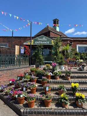 A garden center entrance with a sign, potted plants, and a brick building. - Spacious Airy Double Bedroom Overlooking A Park (London)