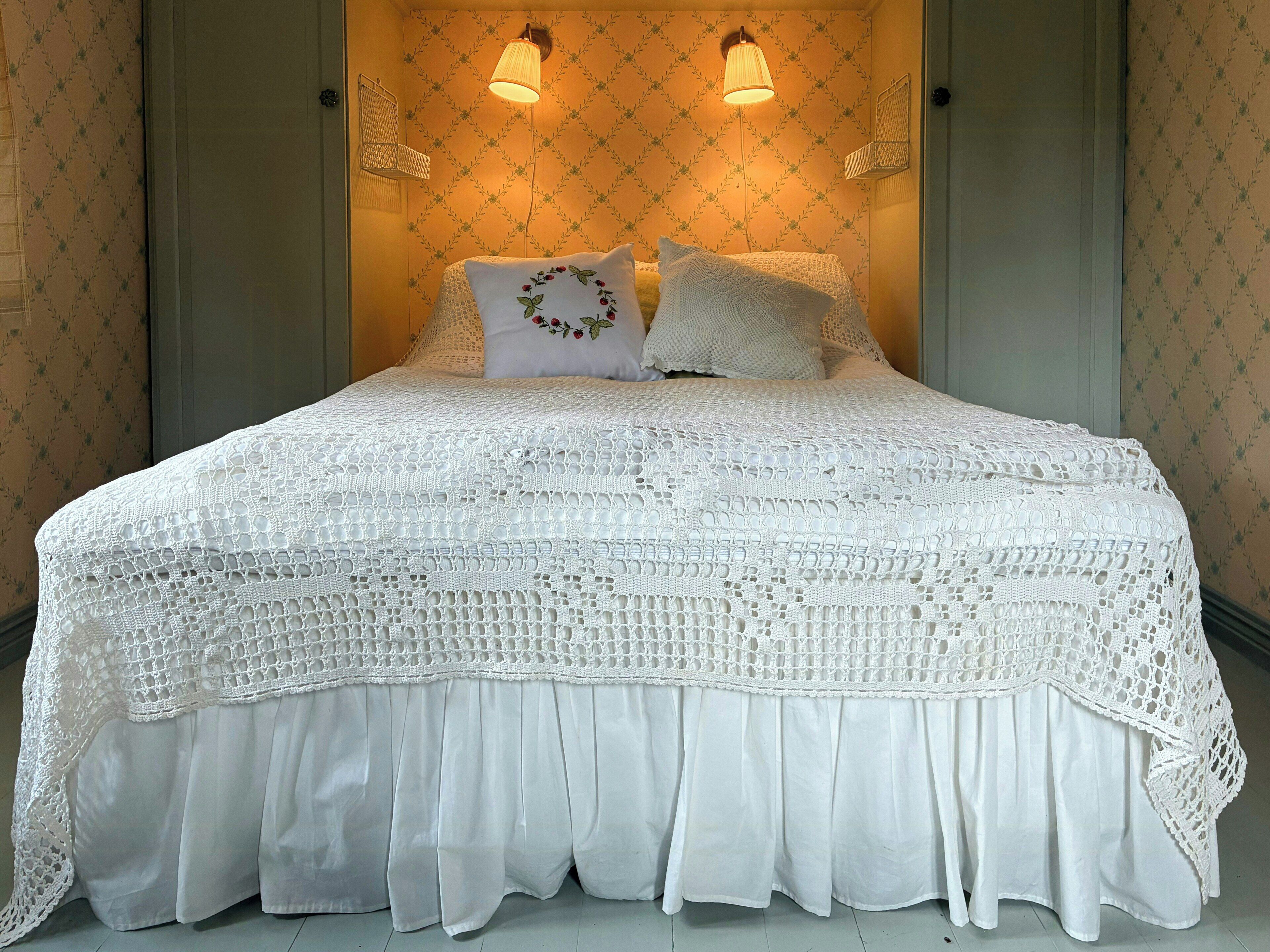Un lit recouvert d’une couvre-lit blanche à motifs complexes, réalisés au crochet, et agrémenté d’oreillers assortis, deux lampes fixées au mur et un papier peint à motifs floraux.