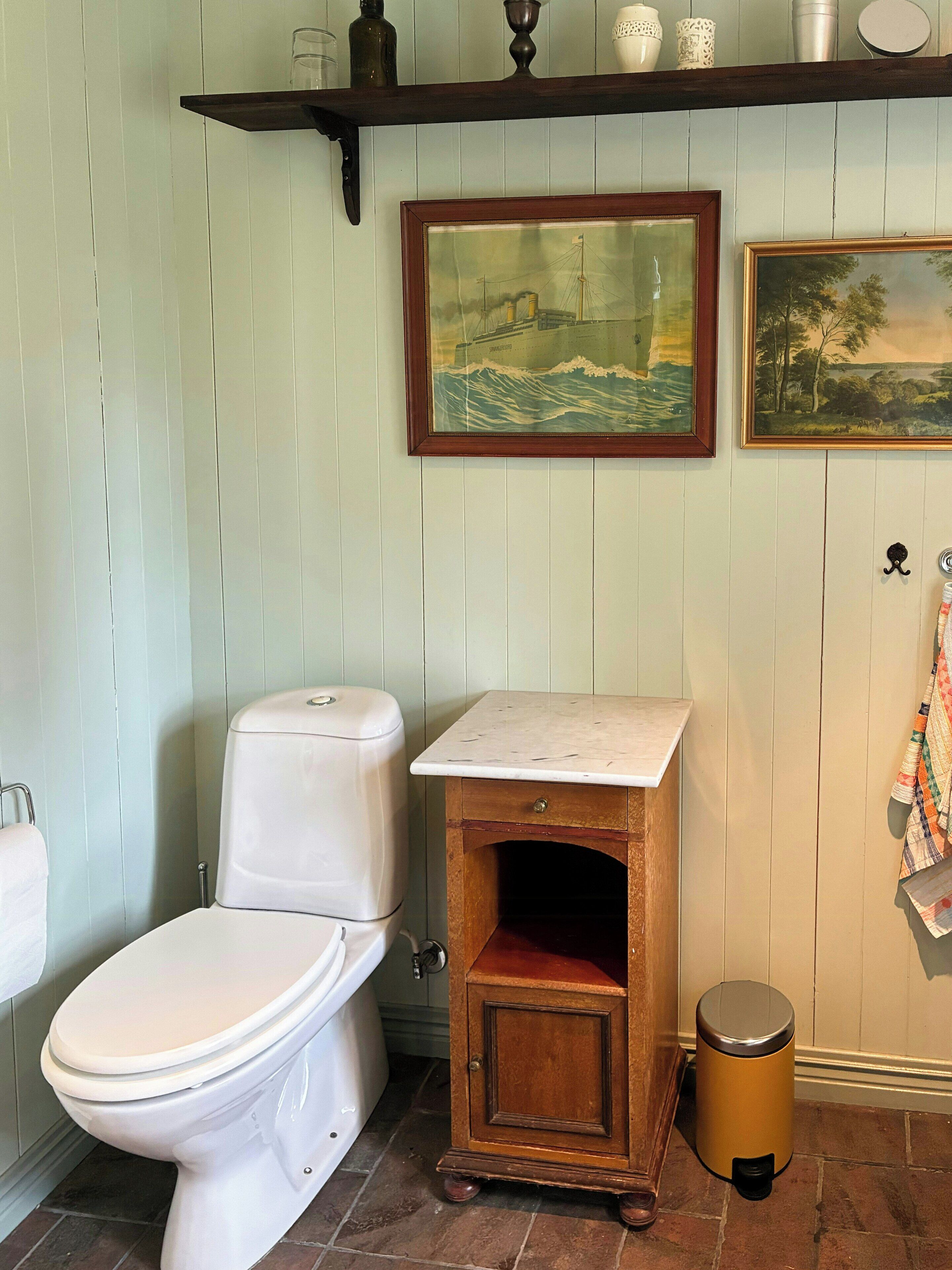 A bathroom with a toilet, a wooden cabinet, a trash can, and two framed pictures on the wall.