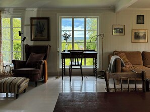 A room with a wooden desk, a chair, a sofa, and a view through large windows.