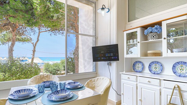 A dining area with a table set for four, a television mounted on the wall, and a view of the sea through large windows. - Tamarè 3 by Casa di Marco (Conchiglie-Alto Lido)