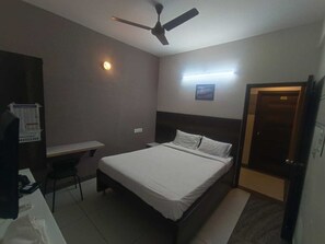 A hotel room with a bed, desk, chair, ceiling fan, and a door.