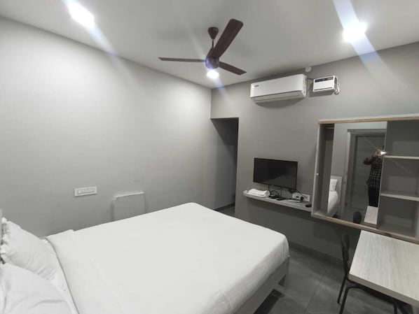 A hotel room with a bed, a desk, a chair, a ceiling fan, an air conditioning unit, a TV, and a mirror.