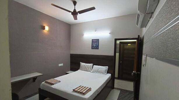 A bedroom with a bed, a ceiling fan, a nightstand, a bedside table, and a door. - Sri Renga Inn (Tiruchirappalli)