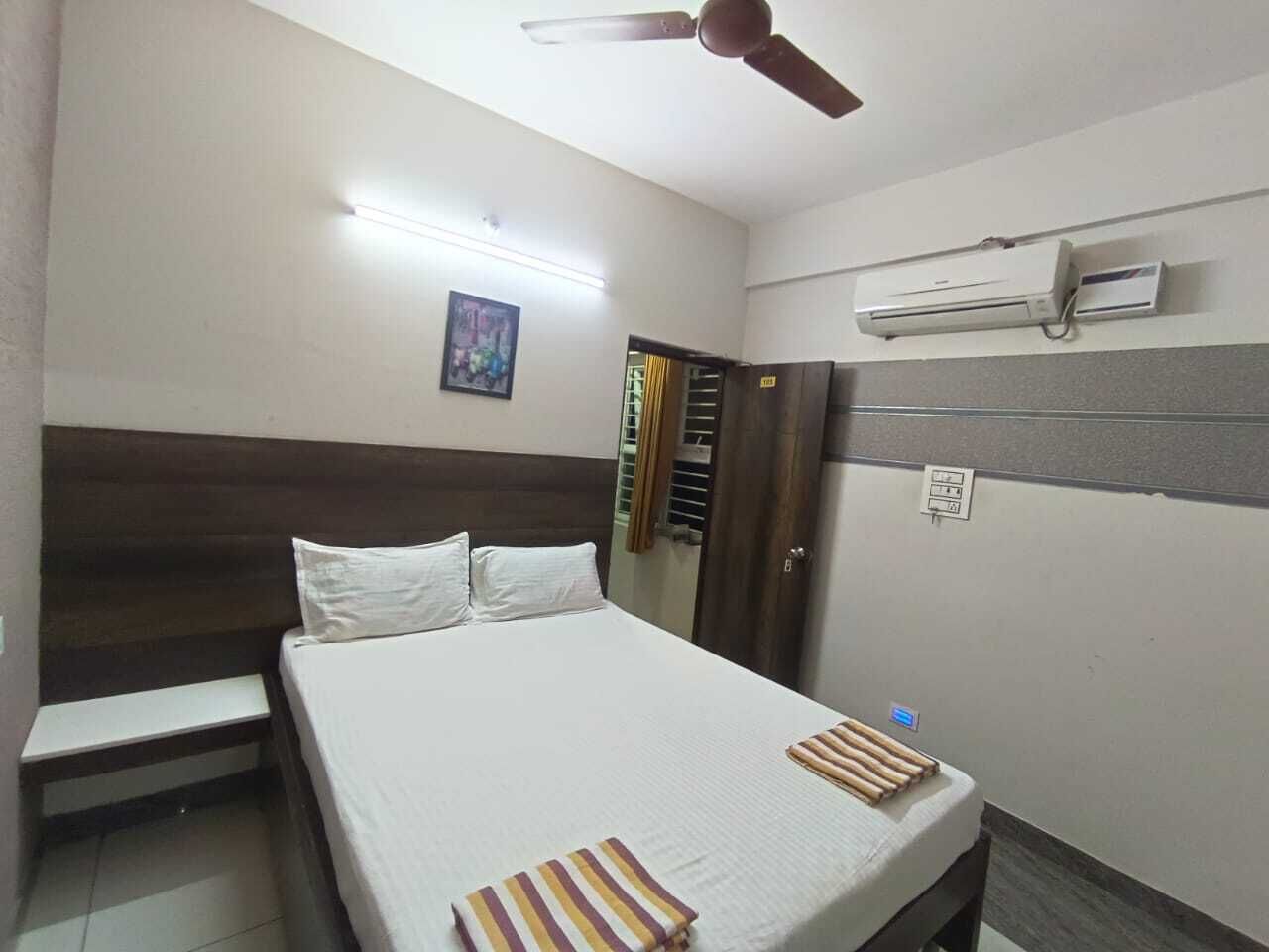 A hotel room with a bed, a ceiling fan, an air conditioning unit, a door with a curtain, and a small bedside table.