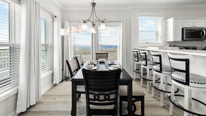 A dining area with a table set for four, surrounded by chairs, a chandelier, and large windows with blinds. - Mariner 1: BeachFront Cottage A, Gulf Front Views! (Mexico Beach)
