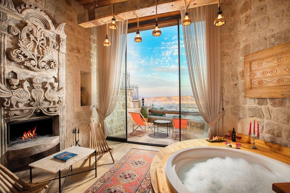 Ein Zimmer mit Stehwänden, einem Kamin, einem großen Fenster mit Vorhängen, einem Holztisch, einer Couch und einer mit Blasen gefüllten Badewanne.
