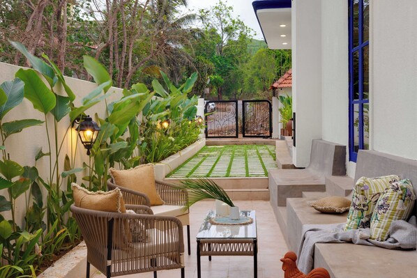 A patio with wicker furniture, a glass table, and a view of a garden and building in the background. - Staymaster Cove Villa - 3BR Private Pool (Parra)