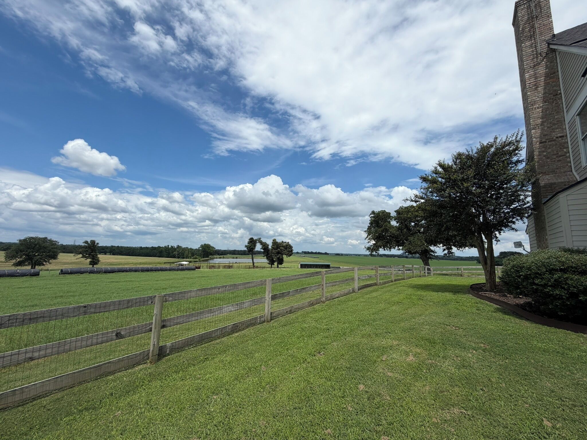 A grassy field with a wooden fence, trees, and a building with a brick chimney.