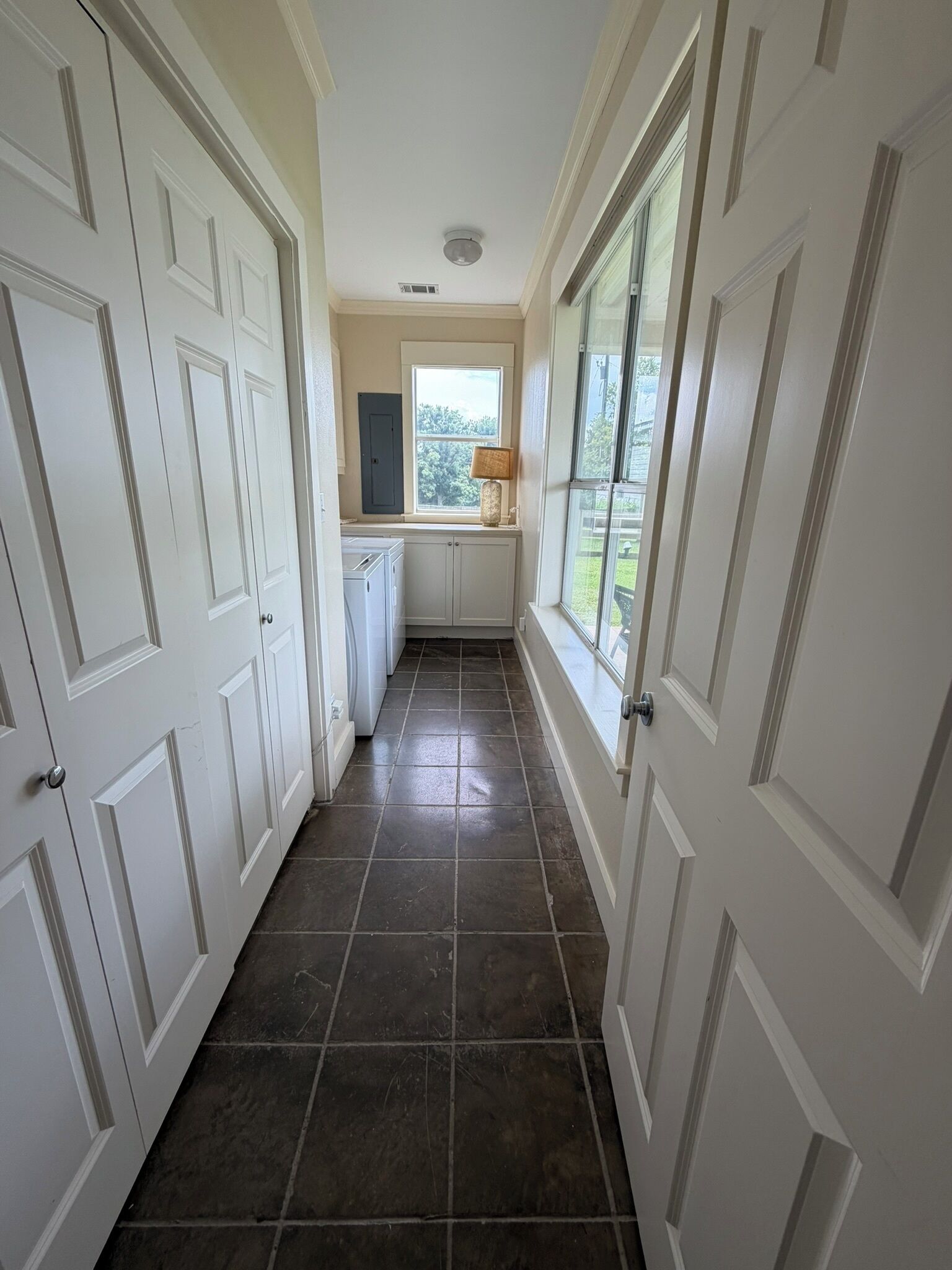 Um corredor com portas brancas, uma janela com vista para o verde e piso de azulejos.