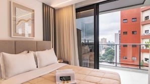 A hotel room with a large bed, a nightstand, a framed picture, and a view of a cityscape through a large window.