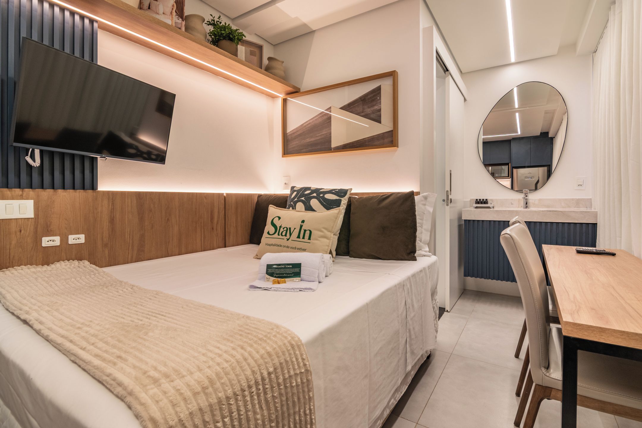 Une chambre d’hôtel moderne équipée d’un lit, d’une table de chevet, d’une télévision, d’un miroir et d’un petit coin repas.