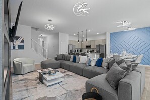Ein modernes Wohnzimmer mit einem grauen Ecksofa, einem weißen Sessel und einem Couchtisch. Im Hintergrund ist eine Küche mit einer blauen Akzentwand zu sehen.