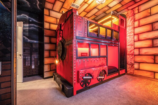 Une chambre aux couleurs vives, avec un thème ferroviaire, un train à deux étages rouge et un mur en briques servant de décor, ainsi qu’une fresque représentant un ciel étoilé.