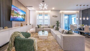 Ein modernes Wohnzimmer mit einem großen Flachbildfernseher, einer Sofagarnitur, einem Couchtisch und einem Essbereich mit Stühlen und Tisch.