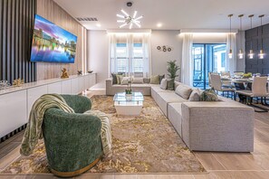 A modern living room with a large flat-screen TV, a sofa set, a coffee table, and a dining area with chairs and a table. - Spacious Luxury: 6BR Condo with Modern Amenities (Orlando)
