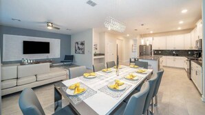 Een moderne keuken met witte kasten, een eethoek met een lange tafel voor zes personen, en een flatscreen televisie aan de wand.