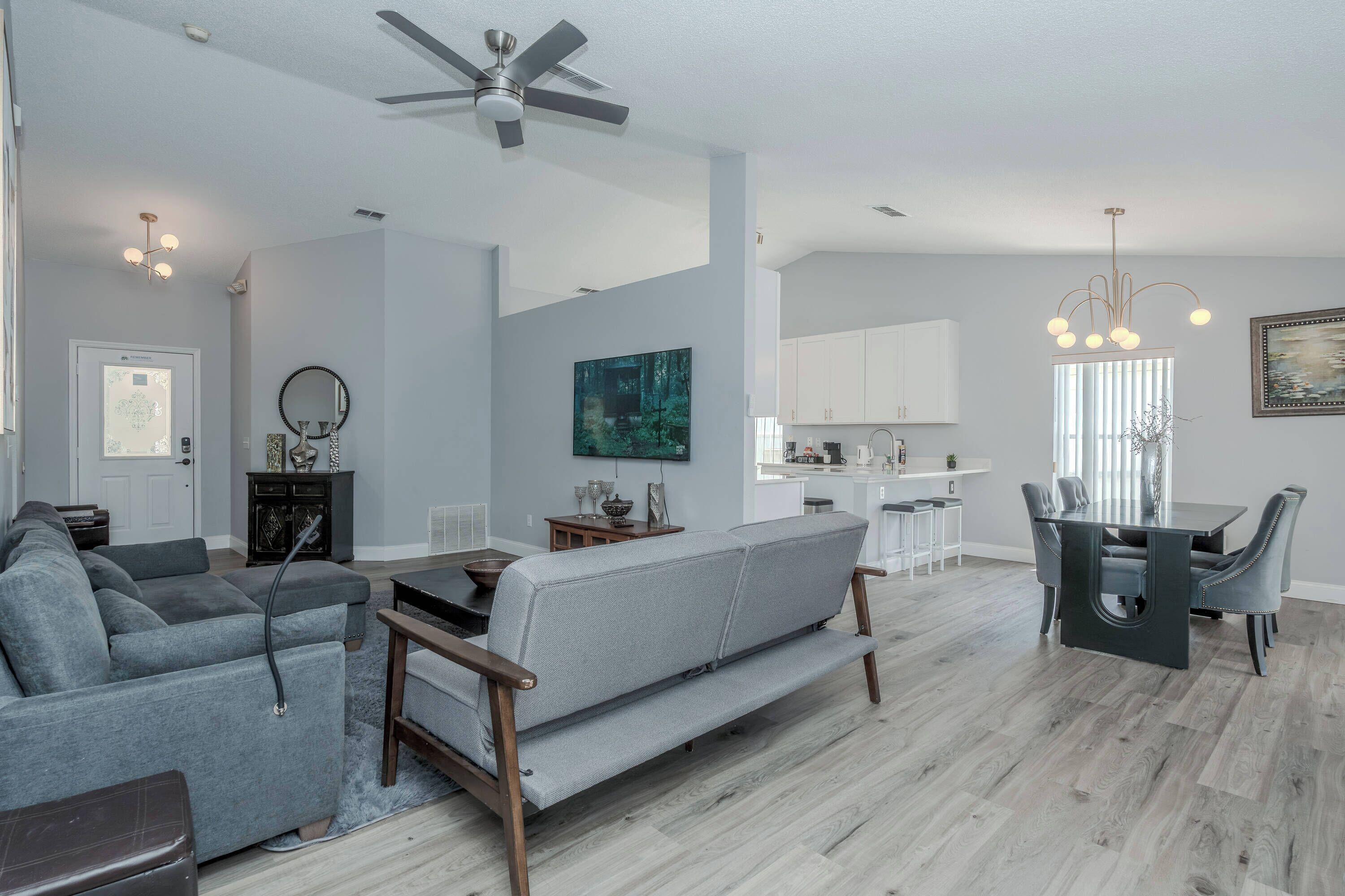 A modern living room with a sectional sofa, a dining area with a table and chairs, a kitchen in the background, and a ceiling fan.