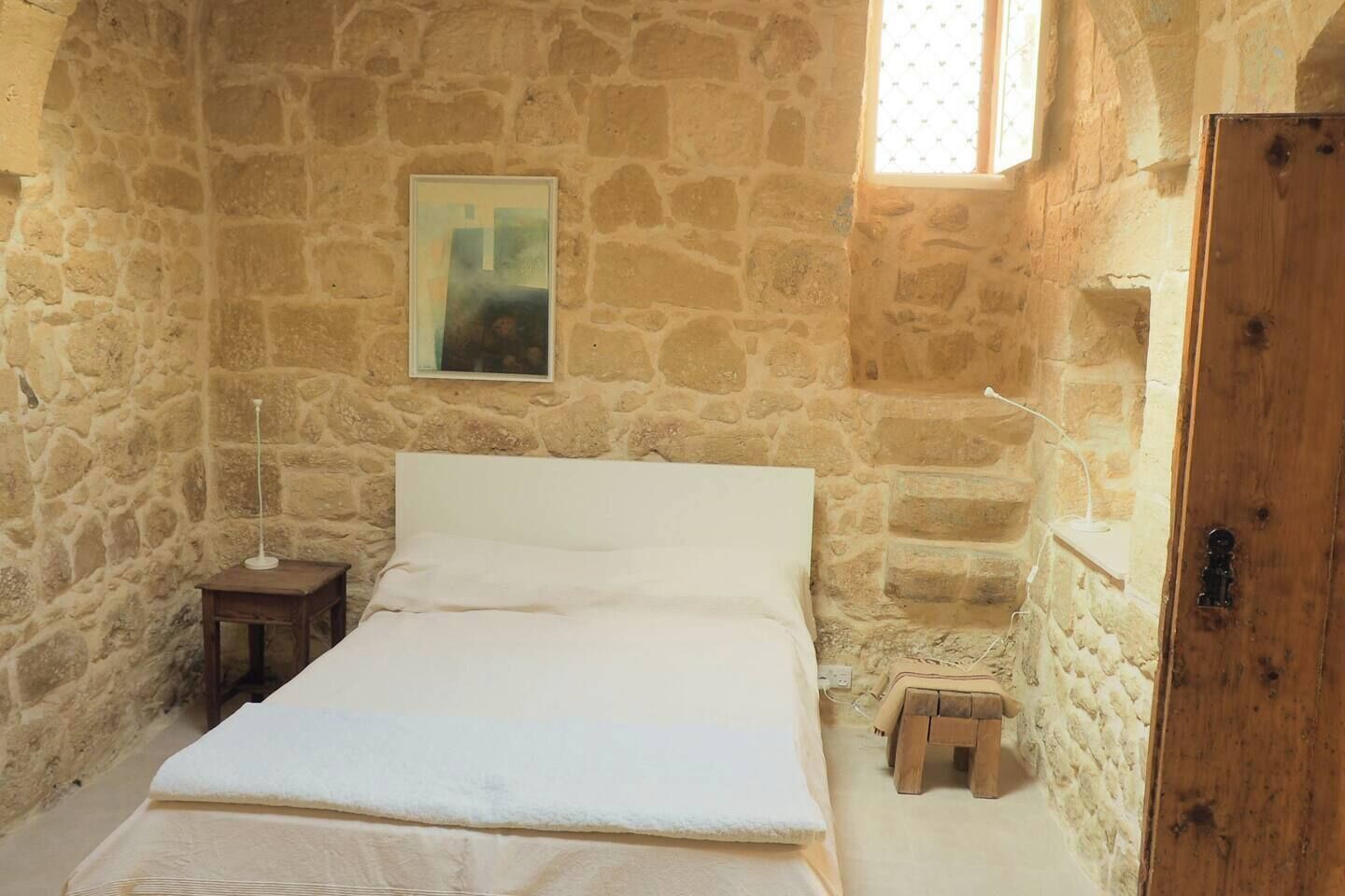 Habitación con paredes de piedra, una cama, una mesita, un taburete y una puerta de madera.