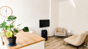 Un salón moderno con una mesa de centro de madera, una planta grande, un sillón color beige, una mesita auxiliar con una lámpara y un televisor montado en la pared.