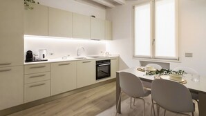 A modern kitchen with white cabinets, a built-in oven, and a dining area with a table and chairs. - GuestHost - Vivaldi Primavera Apartment (Treviglio)