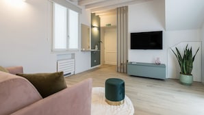 A modern living room with a sofa, a round ottoman, a flat-screen TV, a plant, and a minimalist design. - GuestHost - Vivaldi Primavera Apartment (Treviglio)