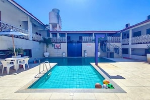 Een zwembad met een basketbalring, omringd door een gebouw met een decoratieve gevel en een heldere lucht.