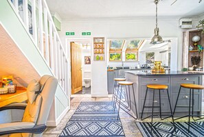 A kitchen with a bar stool, a wooden chair, a patterned rug, and a window with a view. - Hazeldene -  a cottage that sleeps 8 guests  in 3 bedrooms (Shanklin)
