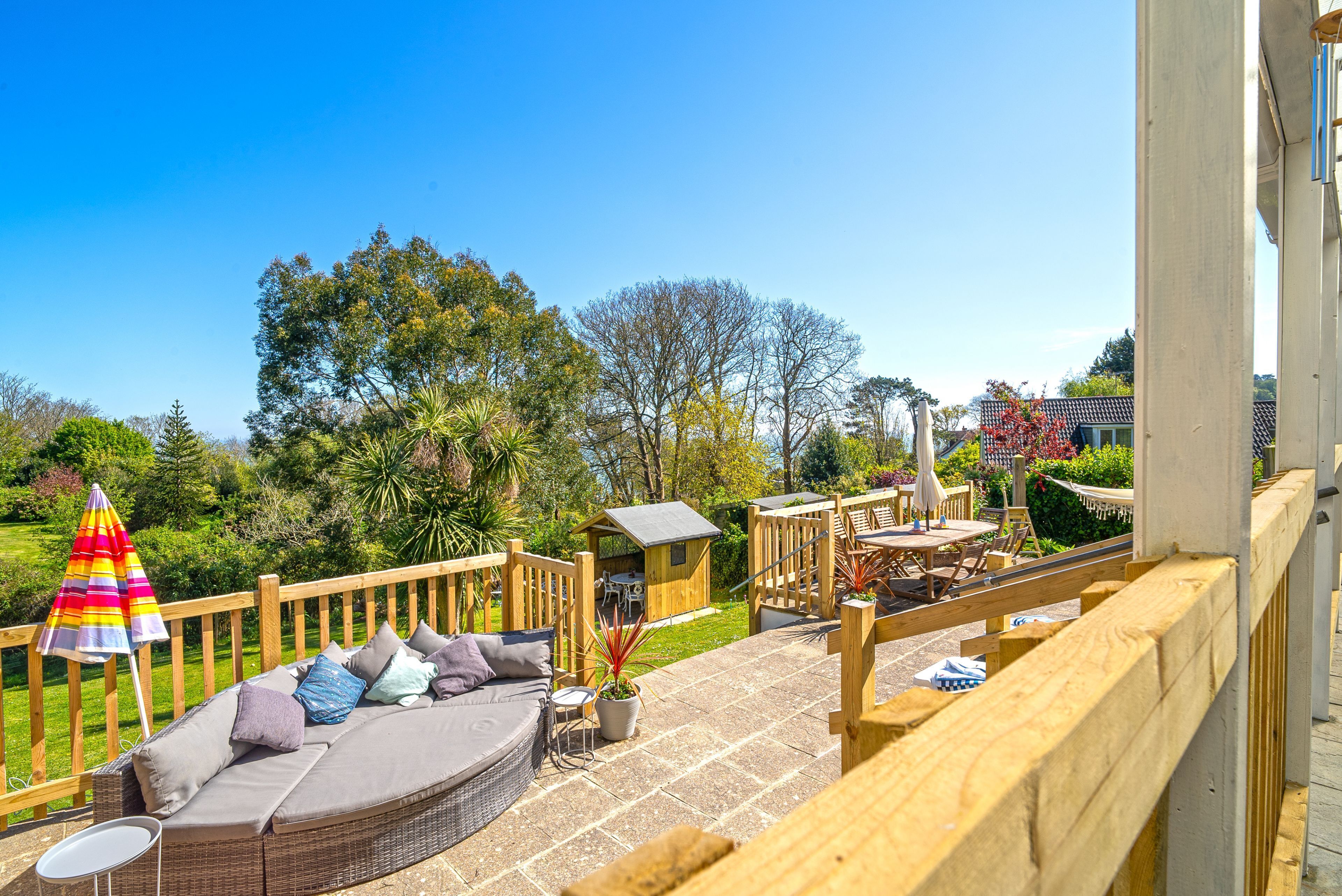 Eine Holzterrasse mit einer Korbsitzgruppe, einem kleinen Tisch und einem bunten Sonnenschirm.