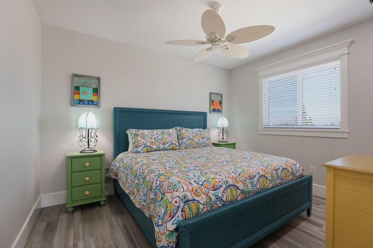 A bedroom with a bed, a ceiling fan, two bedside tables with lamps, a window with blinds, and artwork on the walls.