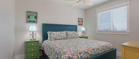 A bedroom with a bed, a ceiling fan, two bedside tables with lamps, a window with blinds, and artwork on the walls.
