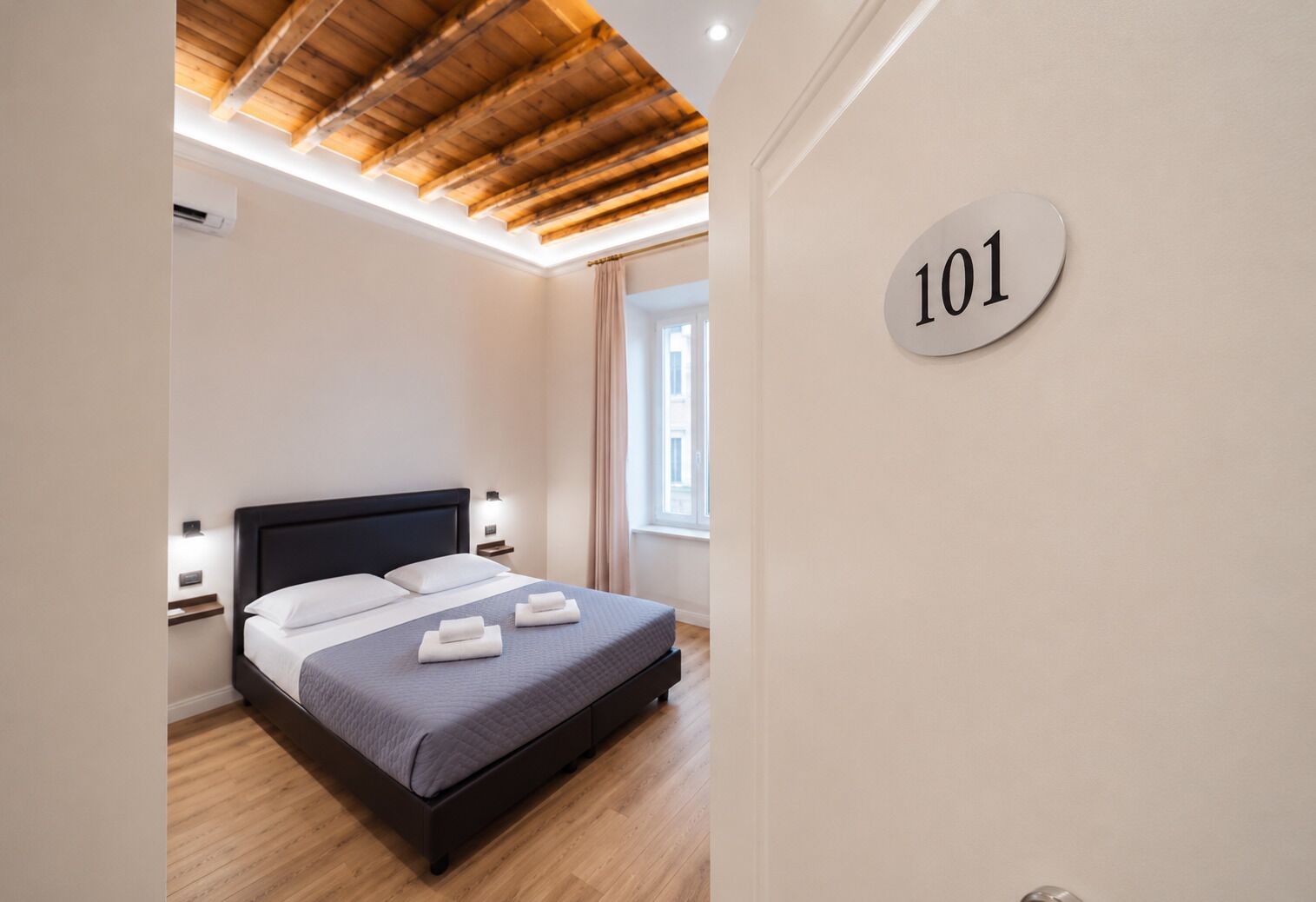 A bedroom with a bed, a headboard, bedside tables, a window, and a wall-mounted sign with the number 101.
