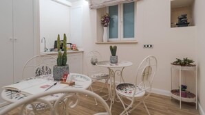 A room with a dining table, chairs, a window with a view, a shelf with decorative items, and a small plant on the windowsill.