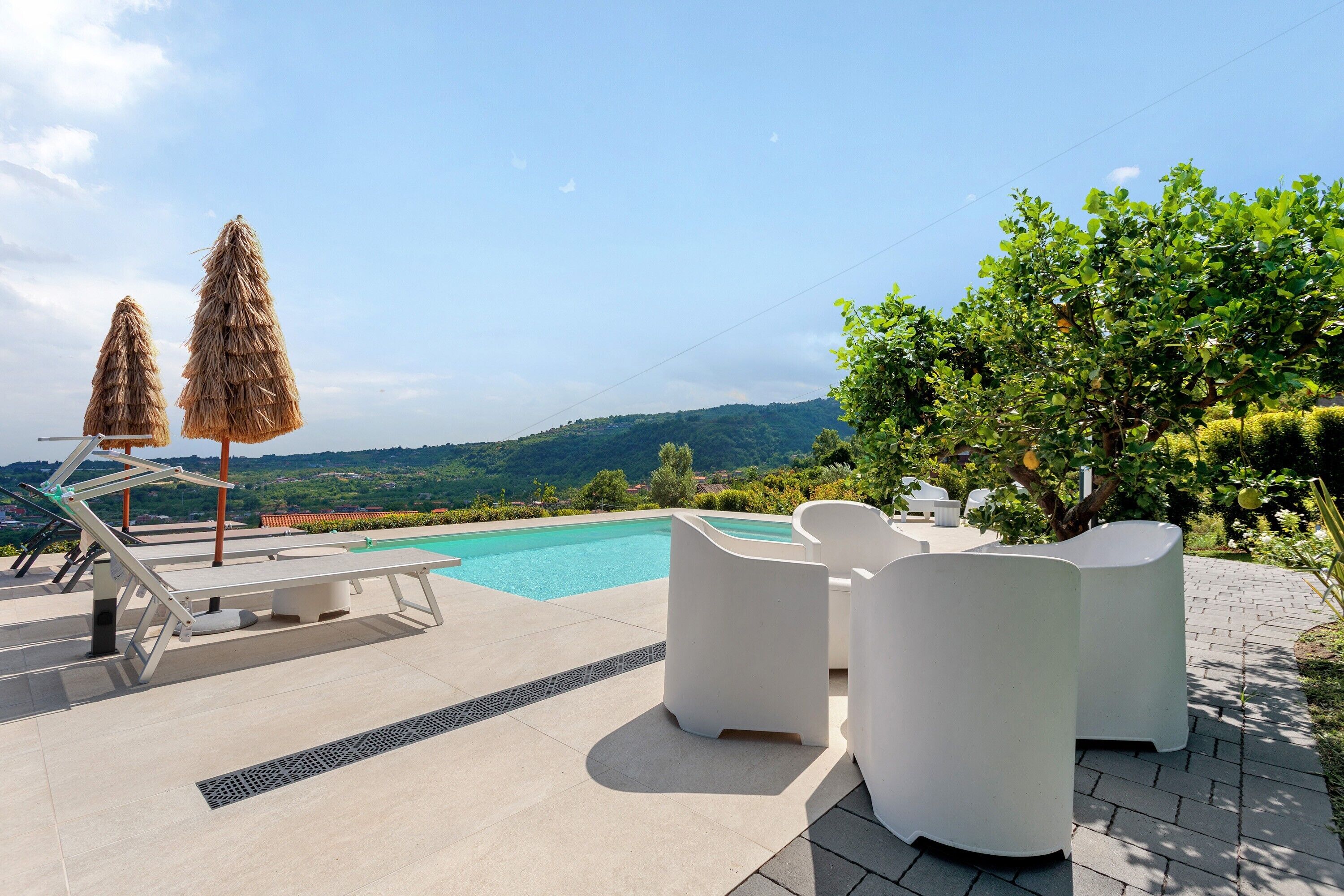 Piscina con sillones de descanso y sombrillas, rodeada de vegetación y colinas.
