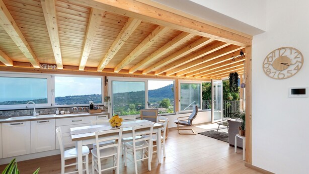 Una cocina moderna con vigas de madera, un comedor con sillas blancas y ventanas grandes que ofrecen vistas al campo.