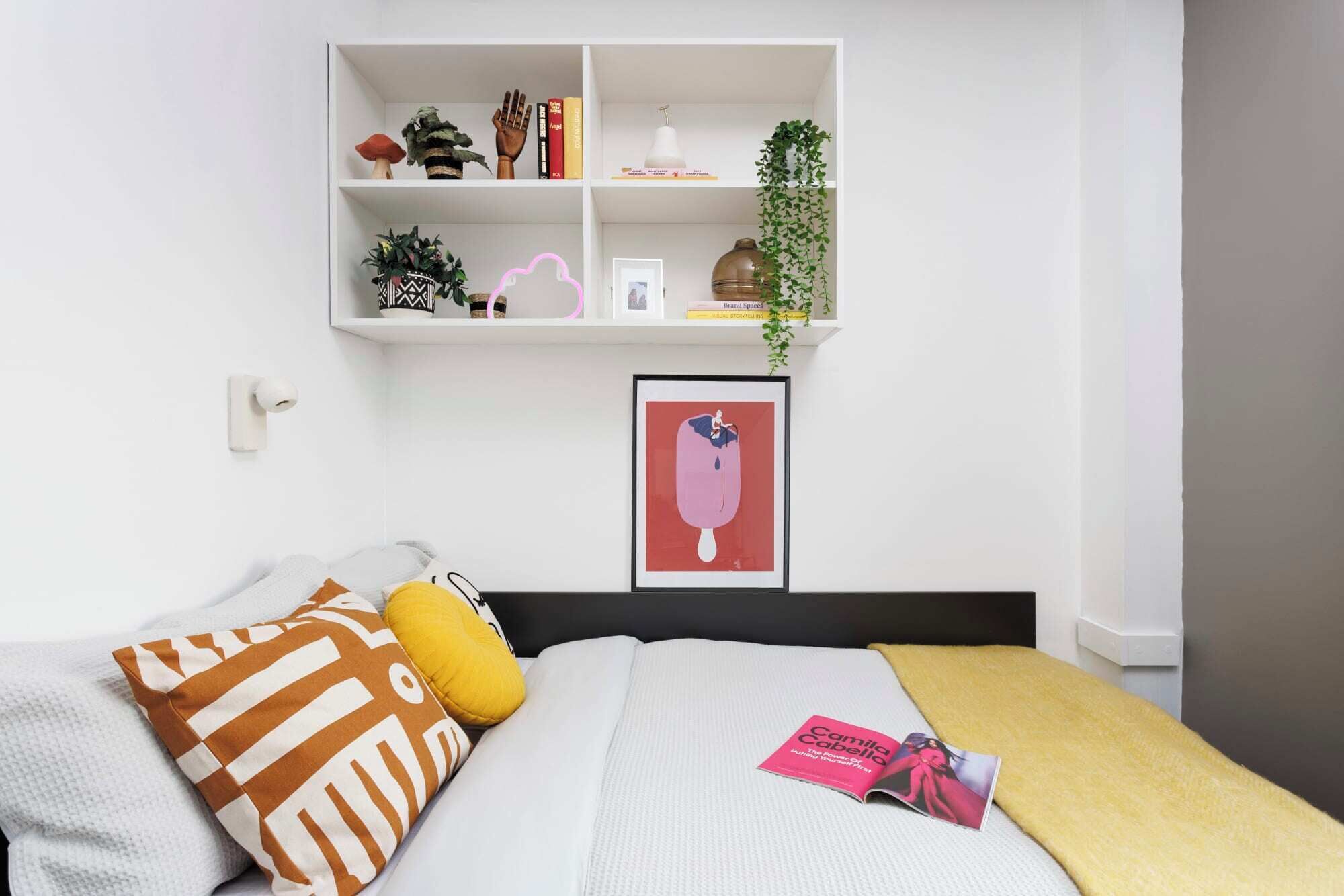 A bedroom with a bed, pillows, a bedside table, a shelf with books and plants, and a framed picture on the wall.