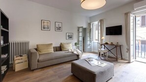 A modern living room with a sofa, ottoman, and a TV. There is a radiator, a bookshelf, and a table with a lamp. - Elegant Apartment with Terrace & Vasari Corridor Views in the Heart of Florence (Firenze)